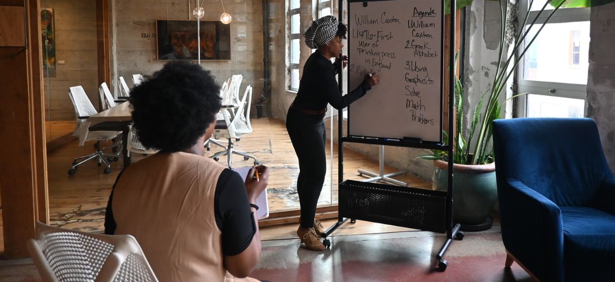 a Black woman writing on a whiteboard for Praxis 2 English tutoring as another woman takes notes